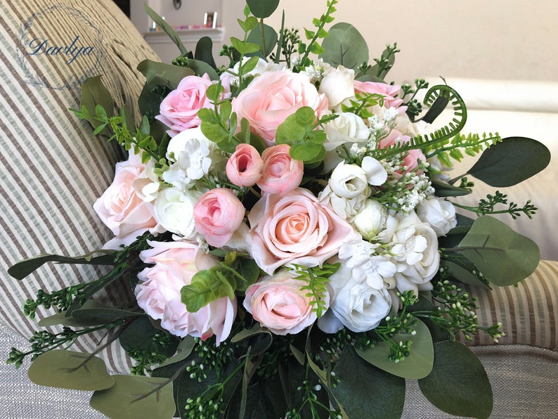 May include: A bridal bouquet with pink and white roses, ranunculus, and greenery. The bouquet is arranged in a classic style and is perfect for a wedding.