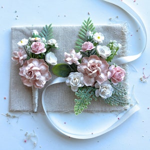 May include: Two pink and white flower bouquets with green leaves on a beige fabric background. The bouquets are tied with a white ribbon.
