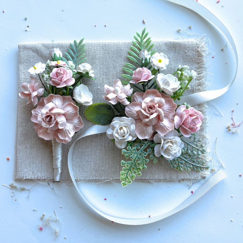 May include: Two pink and white flower bouquets with green leaves on a beige fabric background. The bouquets are tied with a white ribbon.