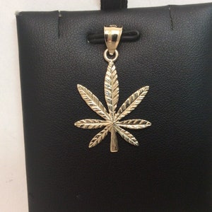 May include: A gold-coloured marijuana leaf pendant charm. The pendant is detailed with a textured design and has a bail for attaching to a chain.