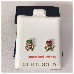 May include: A pair of 14 karat gold frog earrings with green and red gemstones. The earrings are displayed on a white card with the text "Impressed Jewelry 14 KT. GOLD".
