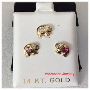 May include: Three gold elephant earrings with clear and pink gemstones. The earrings are displayed on a white card with the text "Impressed Jewelry 14 KT. GOLD".