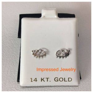 May include: A pair of 14 karat gold ladybug earrings with clear cubic zirconia stones. The earrings are displayed on a white card with the text "Impressed Jewelry 14 KT. GOLD".