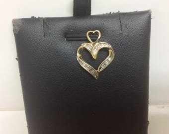 May include: A gold-toned heart pendant with small diamonds set along the curves. The pendant has a small heart at the top and is displayed on a black surface. The jewelry piece is likely a necklace charm or a pendant.