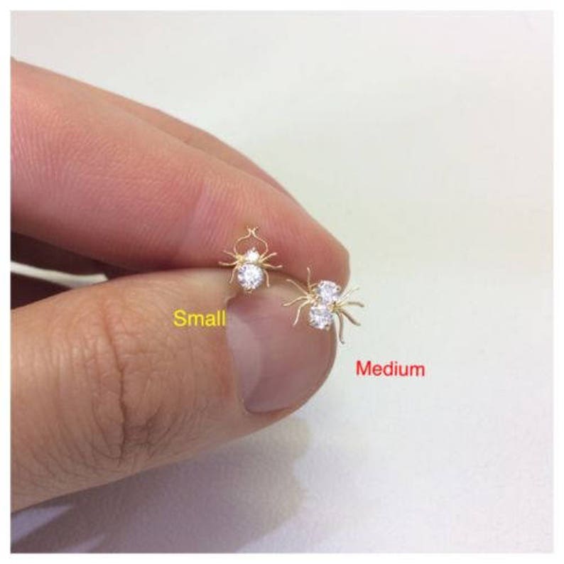 May include: Two pairs of gold spider earrings with clear cubic zirconia stones. The smaller pair is labeled "Small" and the larger pair is labeled "Medium".