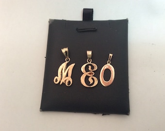 May include: Three gold-colored initial pendants on a black background. The pendants are in the shape of the letters M, E, and O.
