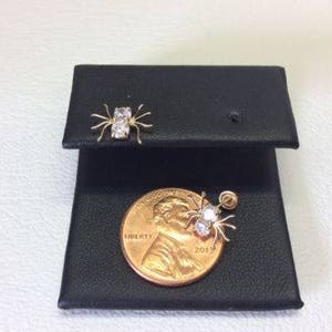 May include: A pair of gold spider earrings with clear cubic zirconia stones. The earrings are displayed on a black jewelry display card with a US penny for scale.