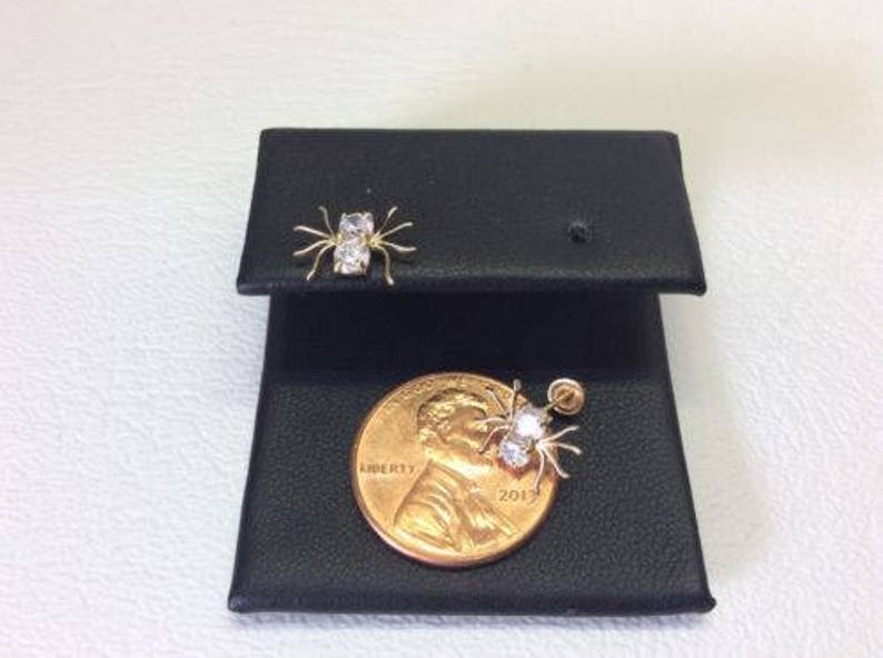 May include: A pair of gold spider earrings with clear cubic zirconia stones. The earrings are displayed on a black jewelry display card with a US penny for scale.