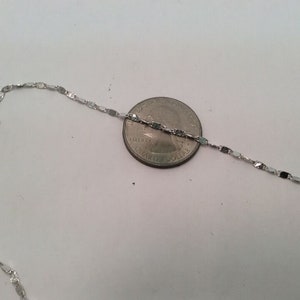 May include: A silver chain necklace with a flat, rectangular link design. The chain is laid out next to a US quarter for size comparison.