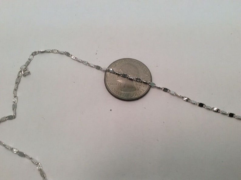 May include: A silver chain necklace with a flat, rectangular link design. The chain is laid out next to a US quarter for size comparison.