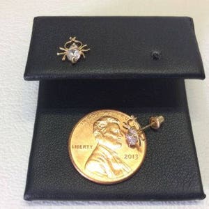 May include: A pair of gold earrings with a small clear stone in the center of each earring. The earrings are displayed on a black surface with a US coin for scale.