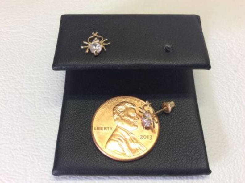 May include: A pair of gold earrings with a small clear stone in the center of each earring. The earrings are displayed on a black surface with a US coin for scale.