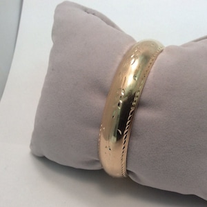 Puede incluir: Un brazalete de oro con un diseño texturizado. El brazalete está sobre un cojín gris.
