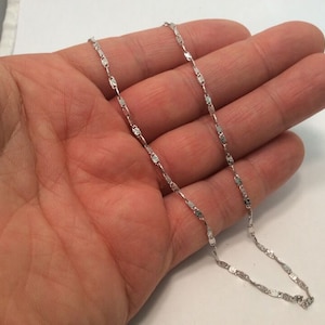 May include: A silver chain necklace with a delicate, flat link design. The chain is shown on a person's hand.