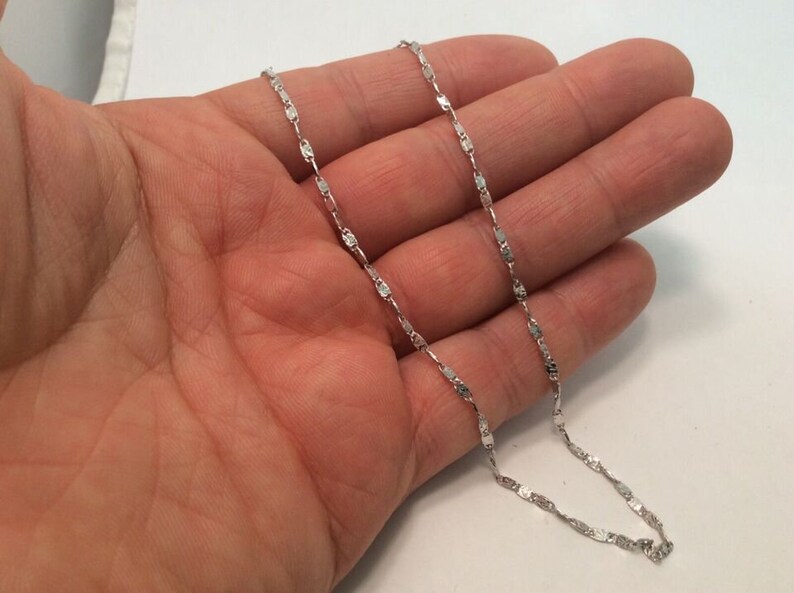 May include: A silver chain necklace with a delicate, flat link design. The chain is shown on a person's hand.