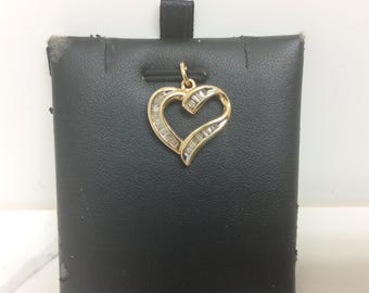 May include: A gold pendant in the shape of a heart, encrusted with small, rectangular-cut diamonds. The pendant hangs from a small loop and is displayed on a black surface. The heart design is open, allowing light to pass through.