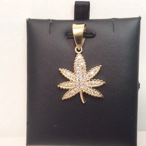 Puede incluir: Un colgante de oro en forma de hoja de marihuana con piedras de circonita cúbica transparentes.