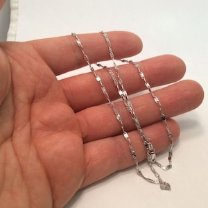 May include: A silver chain necklace with a delicate, flat, and slightly textured design. The chain is layered on a hand, showing the length and detail of the necklace.