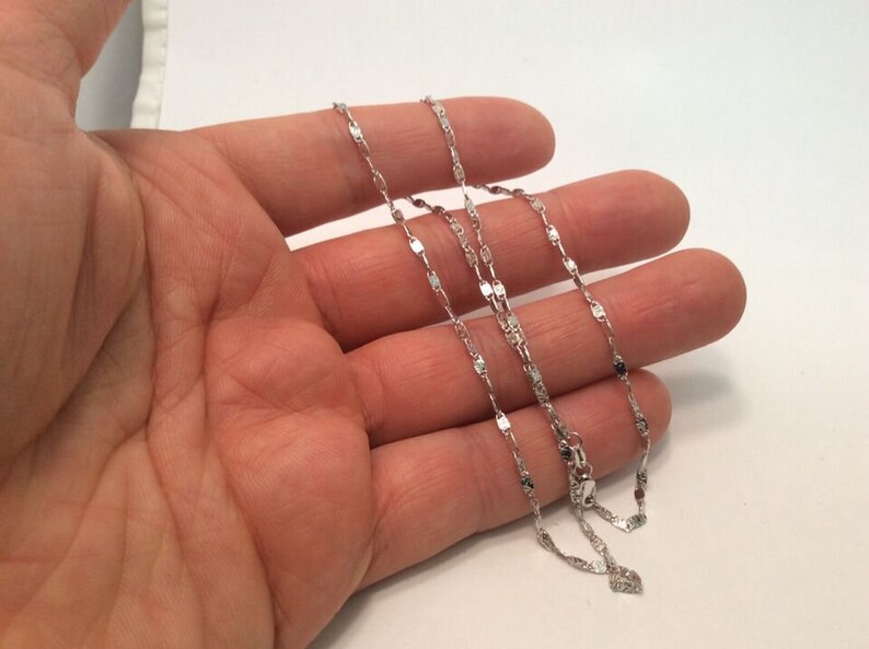 May include: A silver chain necklace with a delicate, flat, and slightly textured design. The chain is layered on a hand, showing the length and detail of the necklace.