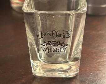 May include: A clear, square shot glass with the words "Jack Daniel's Whiskey" printed in black. The glass has a thick base and is sitting on a wooden surface. Other glasses are visible in the background.