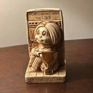 May include: A beige ceramic figurine of a seated child holding a gold trophy. The figurine has the text "MOM IS ANOTHER WORD FOR LOVE" above the child's head. The figurine is on a rectangular base.