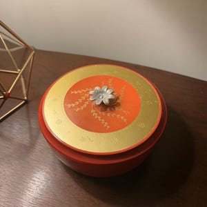 May include: A round, red container with a lid. The lid features a gold border and a central orange circle with gold floral accents. A silver flower-shaped handle is in the center. The container sits on a dark wooden surface.
