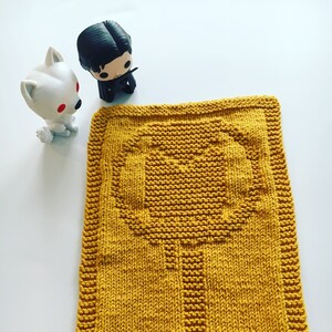 May include: A yellow knitted dishcloth with a tree design. Two small figurines are in the background.