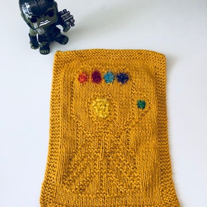 May include: A yellow knitted dishcloth with a design of the Infinity Gauntlet from the Marvel Cinematic Universe. The gauntlet is made with colorful yarn and features six gems.