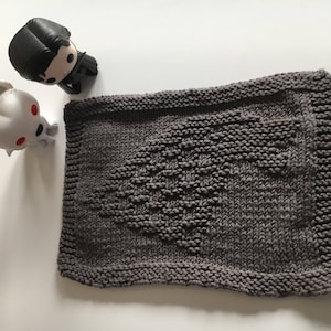 May include: A gray knitted dishcloth with a white, embroidered design of a direwolf. The dishcloth is a square shape with a textured surface.