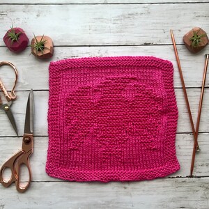 May include: A pink knitted dishcloth with a heart design. The dishcloth is lying on a white wooden surface with two pairs of scissors and two knitting needles.