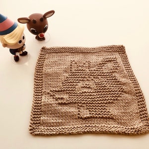 May include: A brown knitted dishcloth with a Christmas tree design. The dishcloth is on a white surface with two small figurines, a reindeer and an elf, in the background.