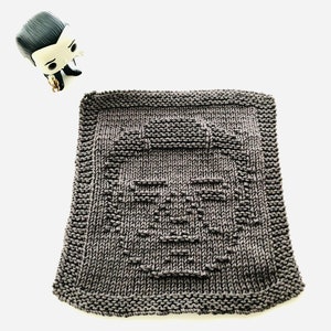 May include: A square, gray knitted washcloth featuring a pixelated face design. The washcloth is made of a soft, textured yarn. A small figurine of a character with black hair and a black suit is in the upper left corner.