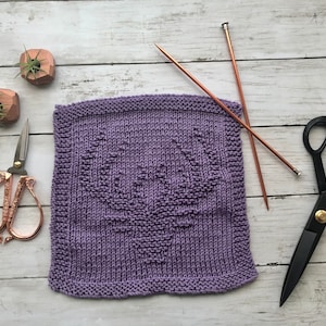 May include: A purple knitted dishcloth with a deer design. The dishcloth is lying on a white wooden surface with two small potted plants and two pairs of scissors.