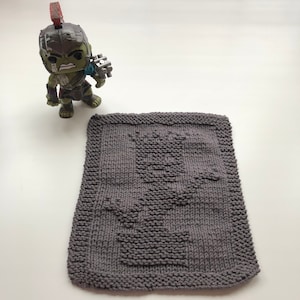 May include: A grey knitted dishcloth with a design of a tree and a pot. The dishcloth is on a white surface.
