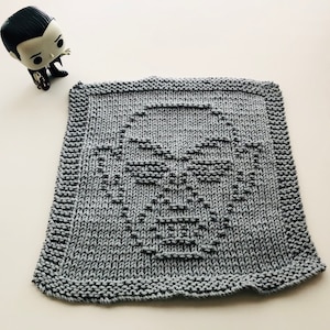 May include: A gray knitted dishcloth with a face design. The face is made with a simple stitch pattern and has a dark gray outline.