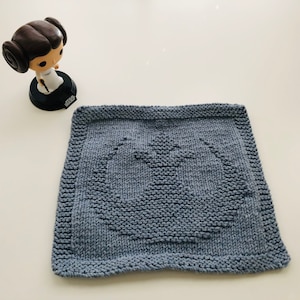 May include: A square, knitted, light blue dishcloth featuring the Rebel Alliance symbol from Star Wars. The dishcloth is made of a textured yarn and has a detailed, raised design. A small figurine of Princess Leia is in the background.