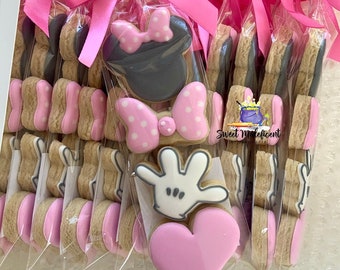 May include: A set of individually packaged cookies decorated with pink and white icing featuring a Minnie Mouse design, a hand with three fingers, and a heart. The cookies are packaged in clear plastic bags tied with pink ribbon.