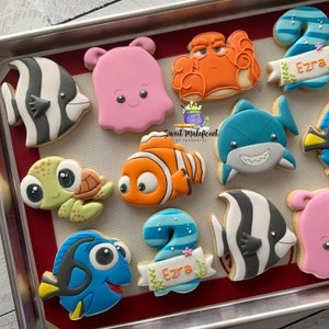 May include: A set of 10 decorated sugar cookies featuring characters from the movie Finding Nemo. The cookies are decorated with colorful icing and include characters like Dory, Nemo, Marlin, Crush, and a shark. The cookies also include the number 2 and the name Ezra.