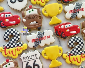 May include: A collection of decorated sugar cookies featuring the characters from the Disney Pixar movie Cars. The cookies are decorated with red, yellow, brown, black, and white icing. The cookies include a red race car, a brown tow truck, a checkered flag, a trophy, and a sign that says "Route 66".