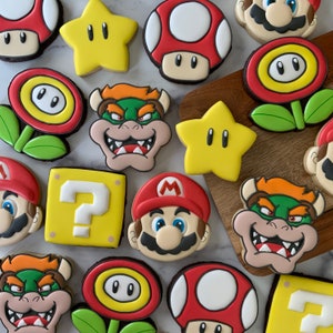 May include: A collection of colorful, decorated sugar cookies featuring various characters and symbols. The cookies are decorated with icing and include characters like Mario, Luigi, and Bowser, as well as symbols like stars and question marks.