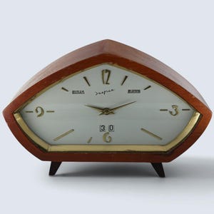 May include: A mid-century style wooden mantel clock with a triangular, shield-shaped frame. The white clock face features gold-toned hands, gold numerals at 3, 6, 9, and 12, and a date display window showing the number 30. It stands on two small feet.