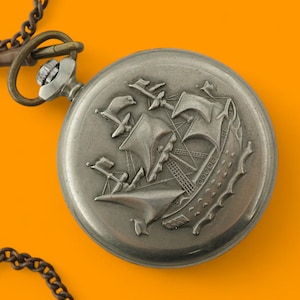 Op de afbeelding: Een zilveren zakhorloge met een gedetailleerde gravure van een zeilschip op de deksel. Het horloge is bevestigd aan een donkere ketting, tegen een heldere oranje achtergrond. Het ontwerp van het horloge suggereert een nautisch of vintage thema.