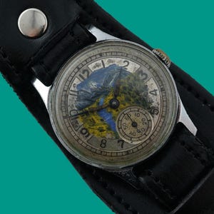 Puede incluir: Reloj de pulsera vintage con caja plateada y correa de cuero negra. La esfera del reloj presenta un diseño de paisaje con una escena de montaña y río. Tiene un pequeño dial de segundos y una corona plateada.