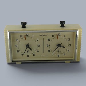 Peut inclure: Une horloge d'échecs analogique rectangulaire de couleur crème avec deux cadrans circulaires, des aiguilles noires et des chiffres noirs. L'appareil dispose de deux boutons-poussoirs noirs sur le dessus et d'un cadre argenté entourant les cadrans. Le texte "Jantar" et "MADE IN USSR" est imprimé sur le cadran.
