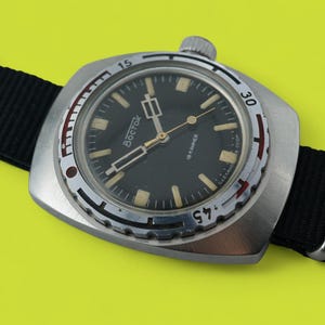 Puede incluir: Un reloj Vostok vintage con esfera negra y caja plateada, con marcadores y agujas luminosas. El reloj está montado en una correa de nailon negro con herrajes plateados. La esfera del reloj muestra la marca "Bostok".