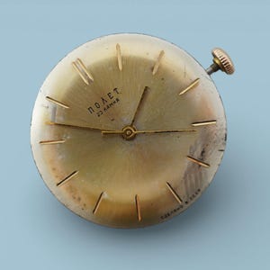 May include: A vintage gold-coloured wristwatch with a round face and a small crown. The watch face features gold-coloured hands and markers, with the text "ПОЛЕТ" and "23 камня" visible. The watch is set against a light blue background.