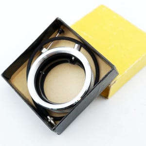 May include: A vintage camera lens adapter kit, presented in its original packaging. The kit includes a silver and black lens adapter ring and a black box with a yellow lid. The items are set against a white background.