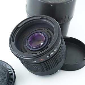 May include: A black camera lens with a purple interior, a lens cap, and a black storage case. The lens has the word "KOHBEPTEP" printed on it. The lens cap is round and black. The storage case is cylindrical and black.