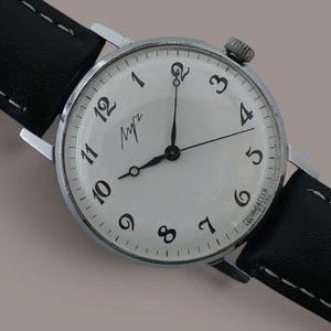 May include: A wristwatch with a round silver-tone case and a white dial featuring black Arabic numerals. The watch has thin black hands and a black leather strap with white contrast stitching. The dial displays the Cyrillic text "Луч" near the top.