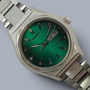 May include: A silver-tone stainless steel wristwatch featuring a round case and a matching link bracelet. The watch has a vibrant emerald green sunburst dial with silver hour markers, silver hands, and a day-date window displaying FRI 14.